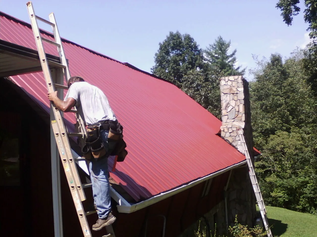 Roofer on a ladder beside a red metal roof and stone chimney — Soffit & Fascia Repair by Roofing Brothers in Farwell